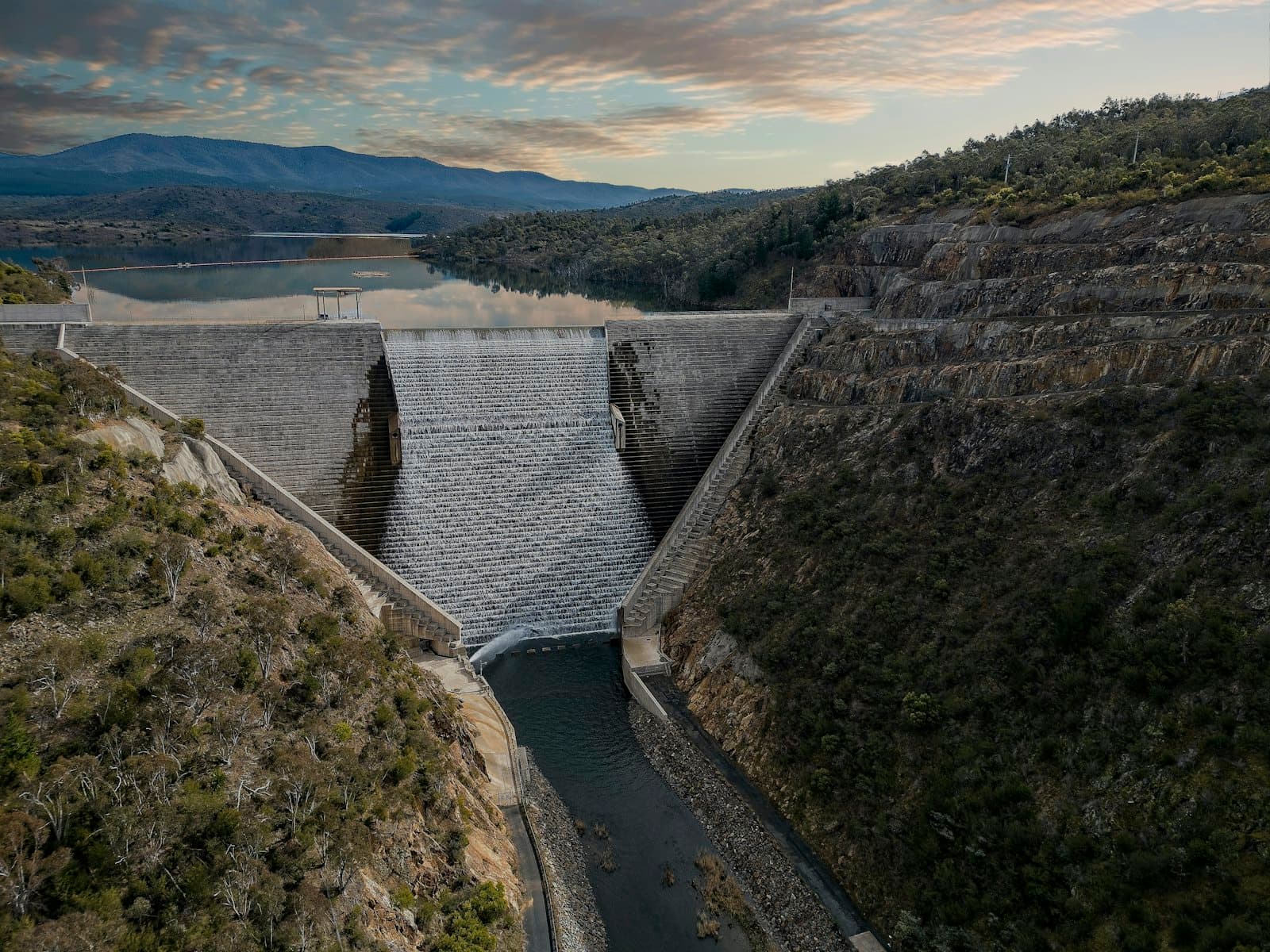 Aerial view of a large dam spillway releasing water into the river below, set against a mountain reservoir